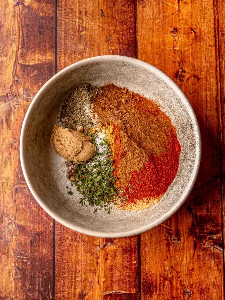 Unmixed jerk seasoning spices in a large mixing bowl, including brown sugar, dried thyme, smoked paprika, and cayenne.
