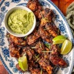 Platter of smoked jerk chicken wings with lime wedges and green dipping sauce in a round bowl.