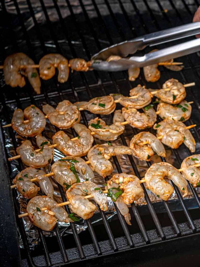 Shrimp skewers being placed on a smoker grate using metal tongs.