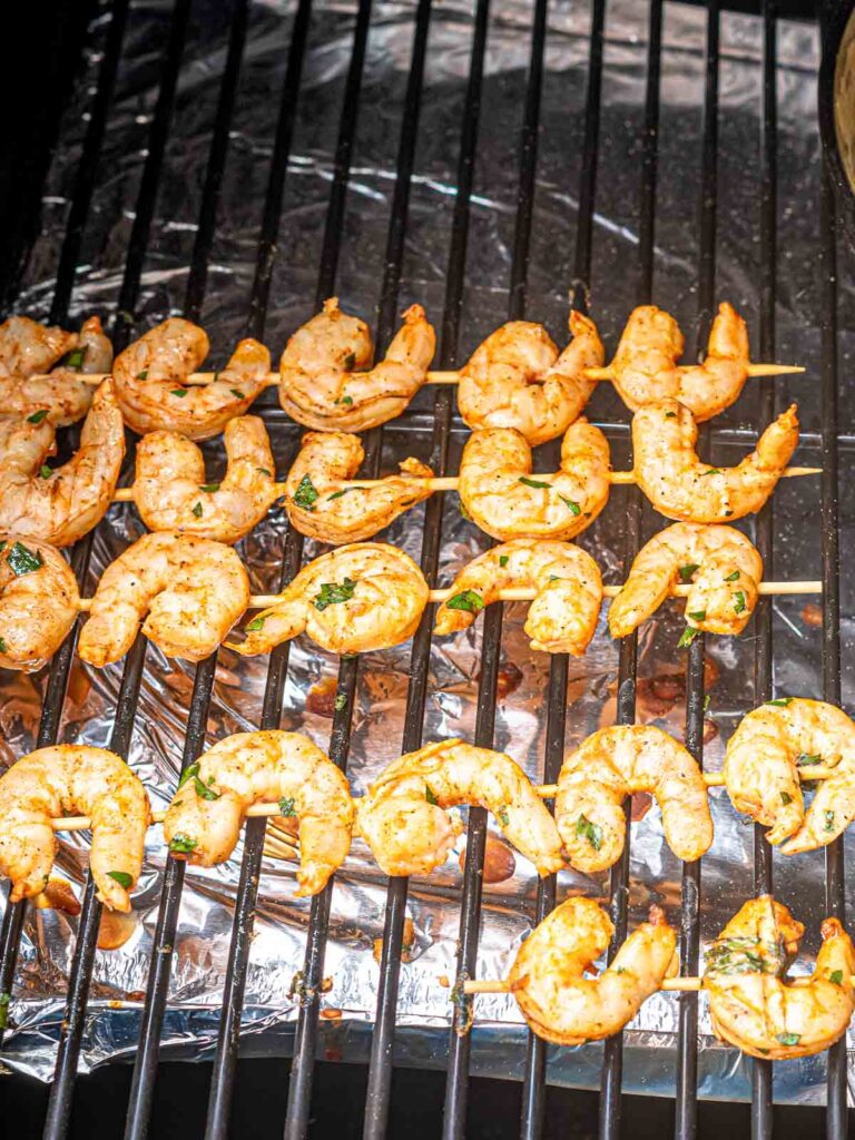 Marinated shrimp skewers smoking on a grill grate lined with foil, halfway through cooking.