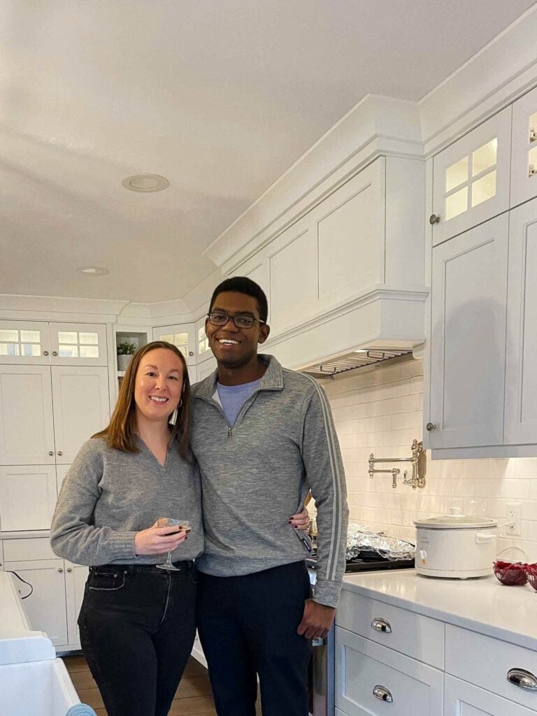 Sarah and Kadeem smiling together in a cozy white kitchen, dressed in coordinating gray tops, holding drinks and standing near a slow cooker on the counter.