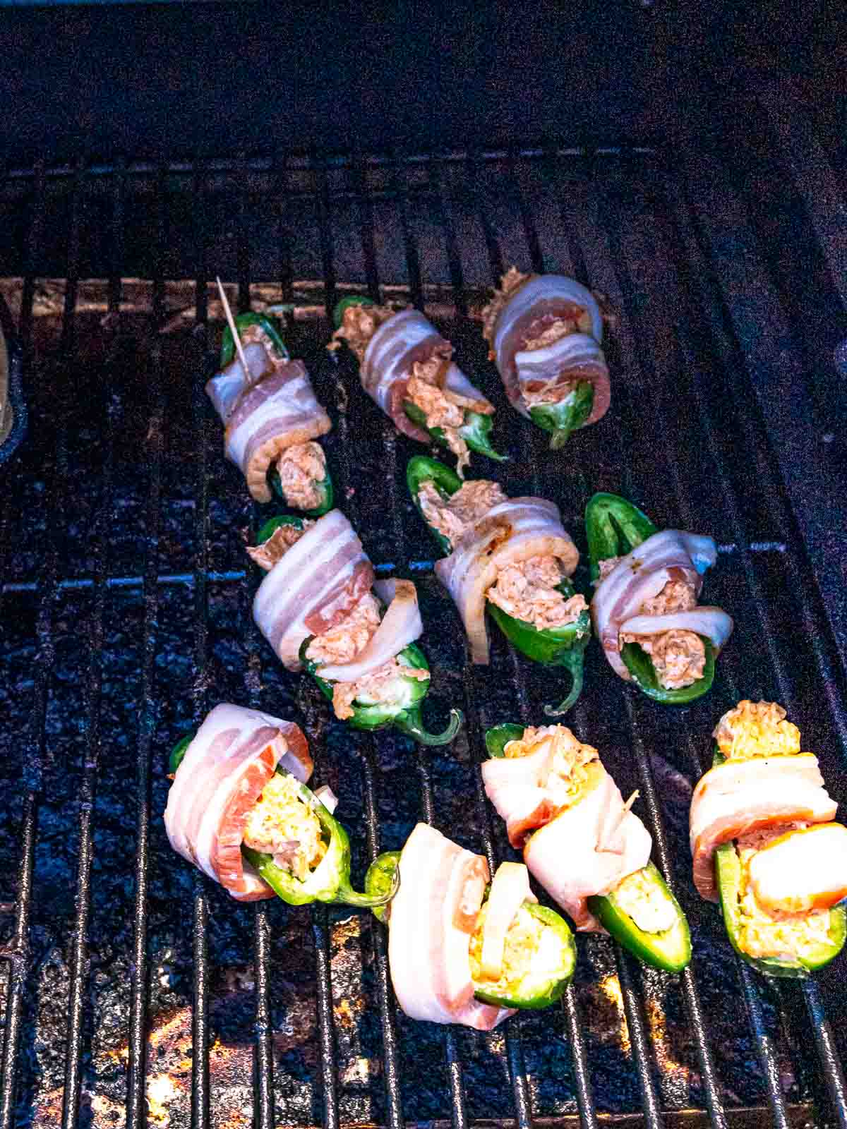 Bacon-wrapped jalapeño poppers cooking on a smoker grate as the bacon begins to render.