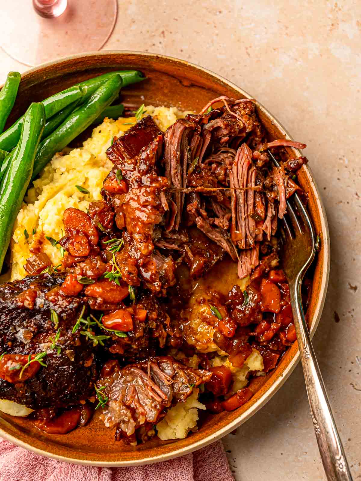 Tender smoked red wine short ribs pulled apart on a plate with mashed potatoes, green beans, and red wine sauce.