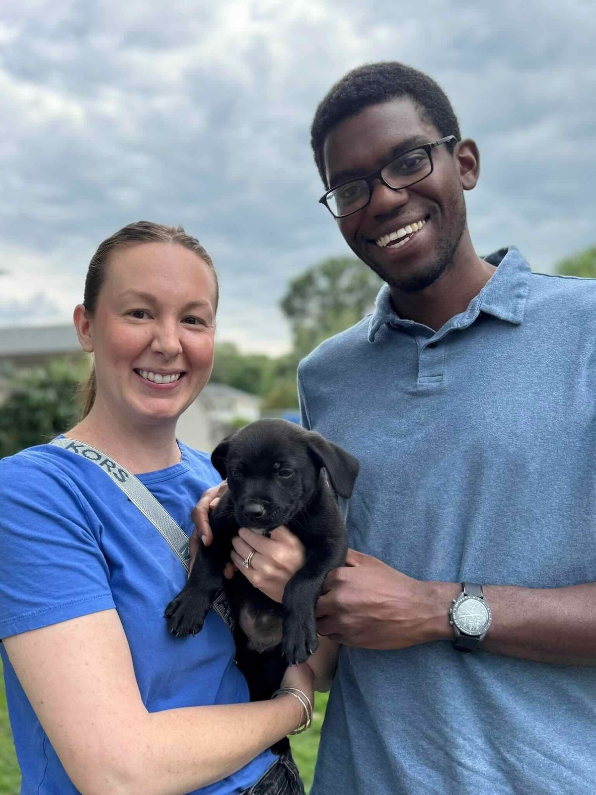 Sarah and Kadeem smiling outdoors while holding a small black puppy.
