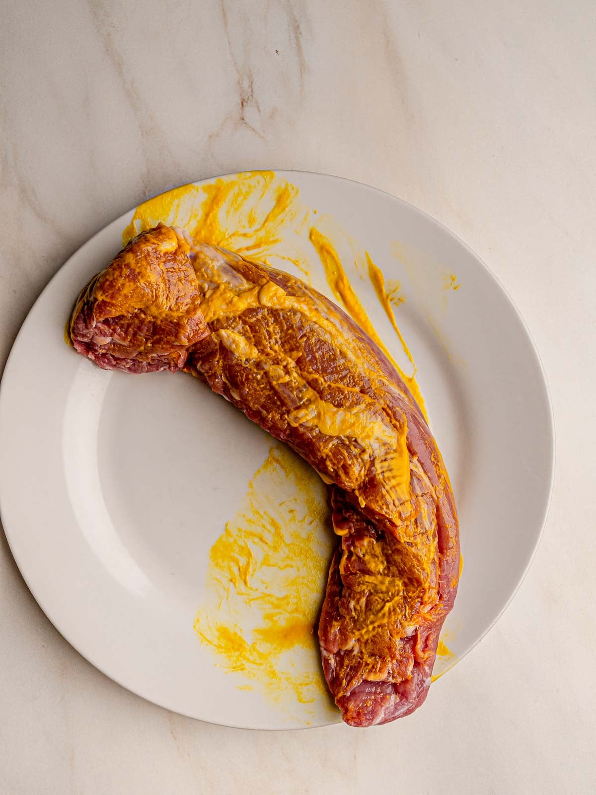 Pork tenderloin coated with yellow mustard on a white plate before seasoning.