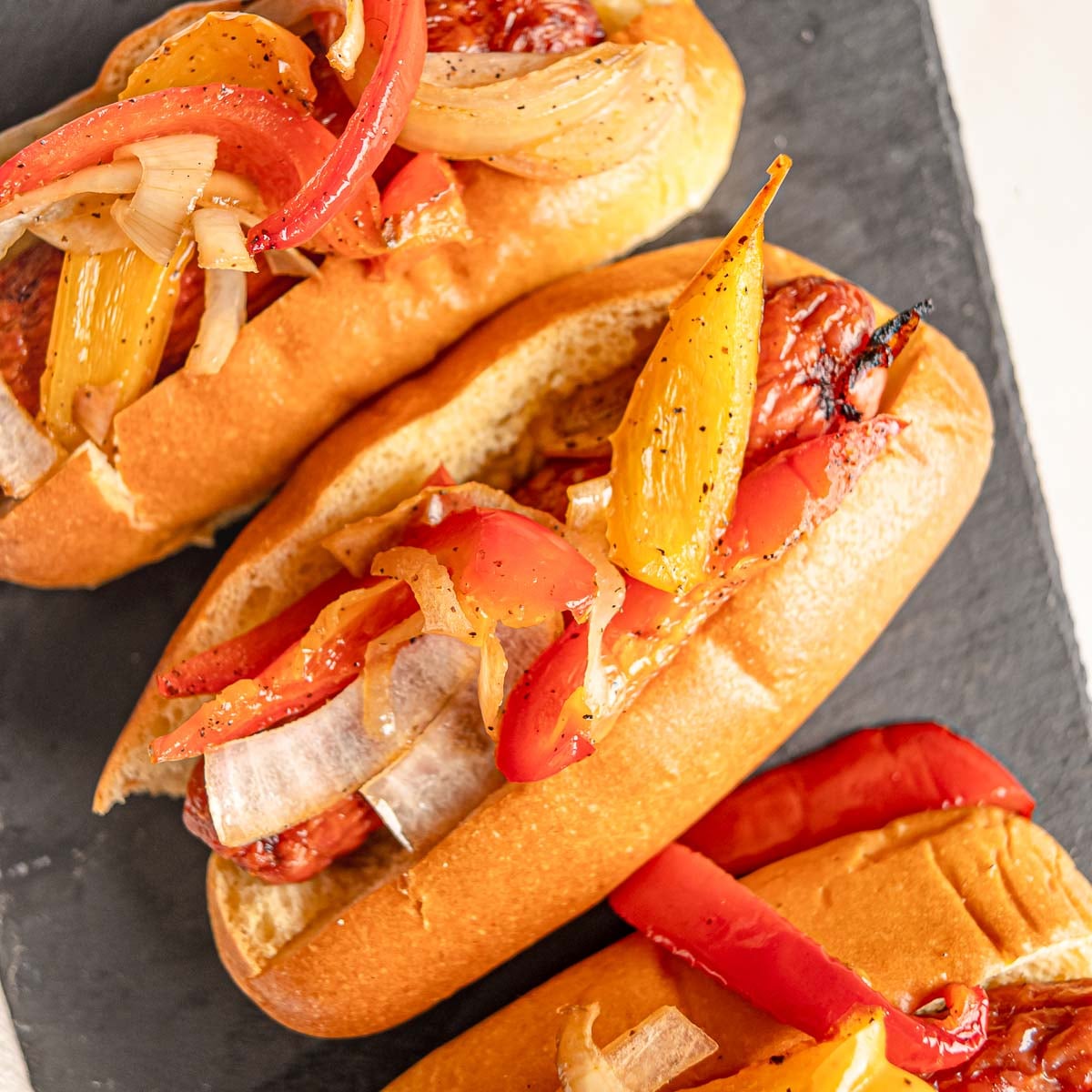 Smoked sausage and peppers served in hoagie rolls on a black slate board.